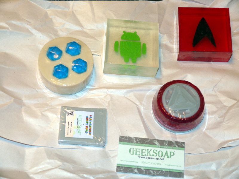 A variety of geeky themed soaps from GeekSoap.net.  From the top left is a round soap with four soap d20 dice embedded in it.  Next is a square soap with the Android logo.  Top right is a square red soap with a Starfleet symbol.  Bottom row left is a small game cartridge soap and bottom right is an Iron Man arc reactor soap.