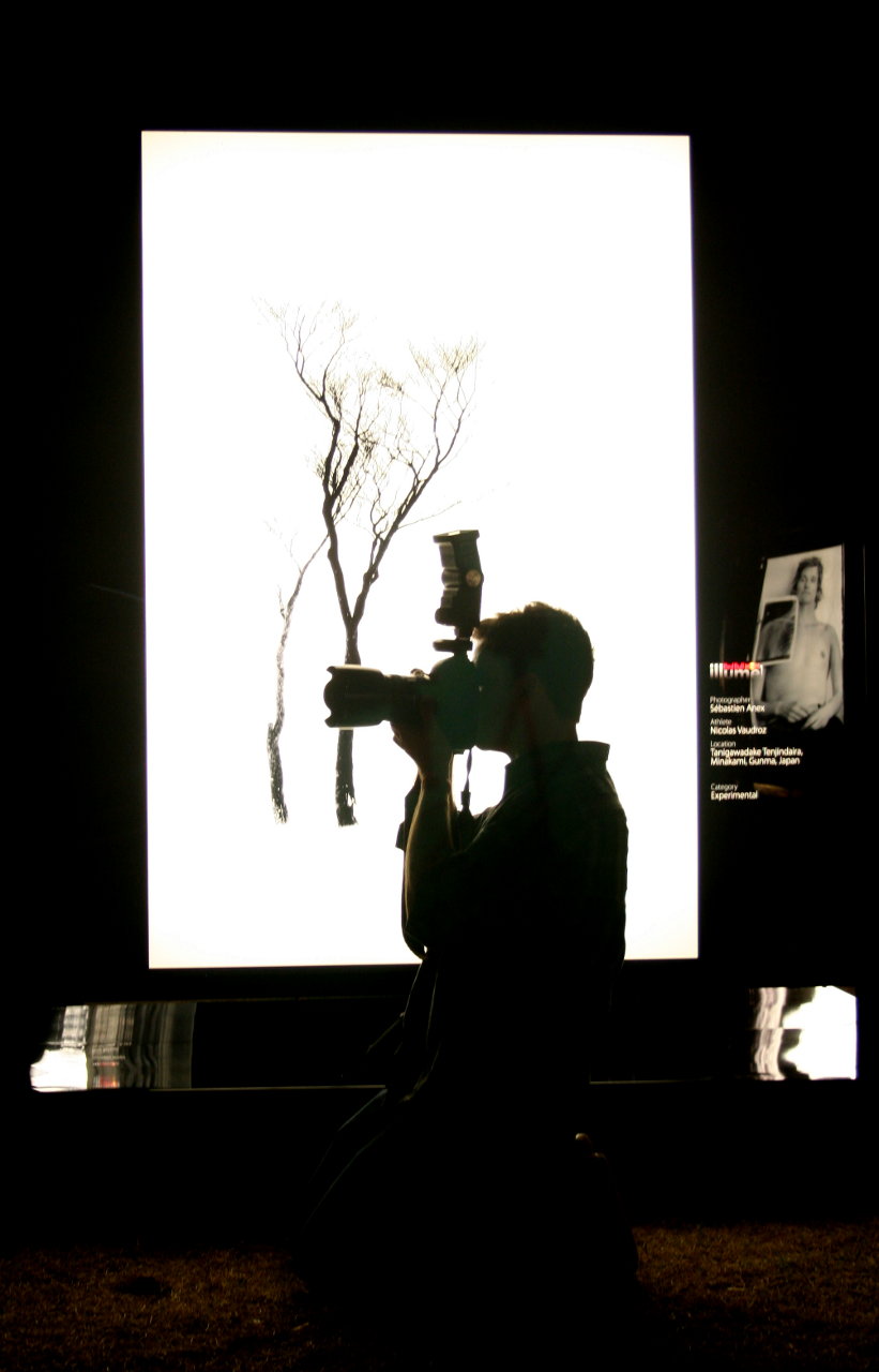 Silhouette of a photographer against the background of an illuminated display.  The display is of a tree against a white background.