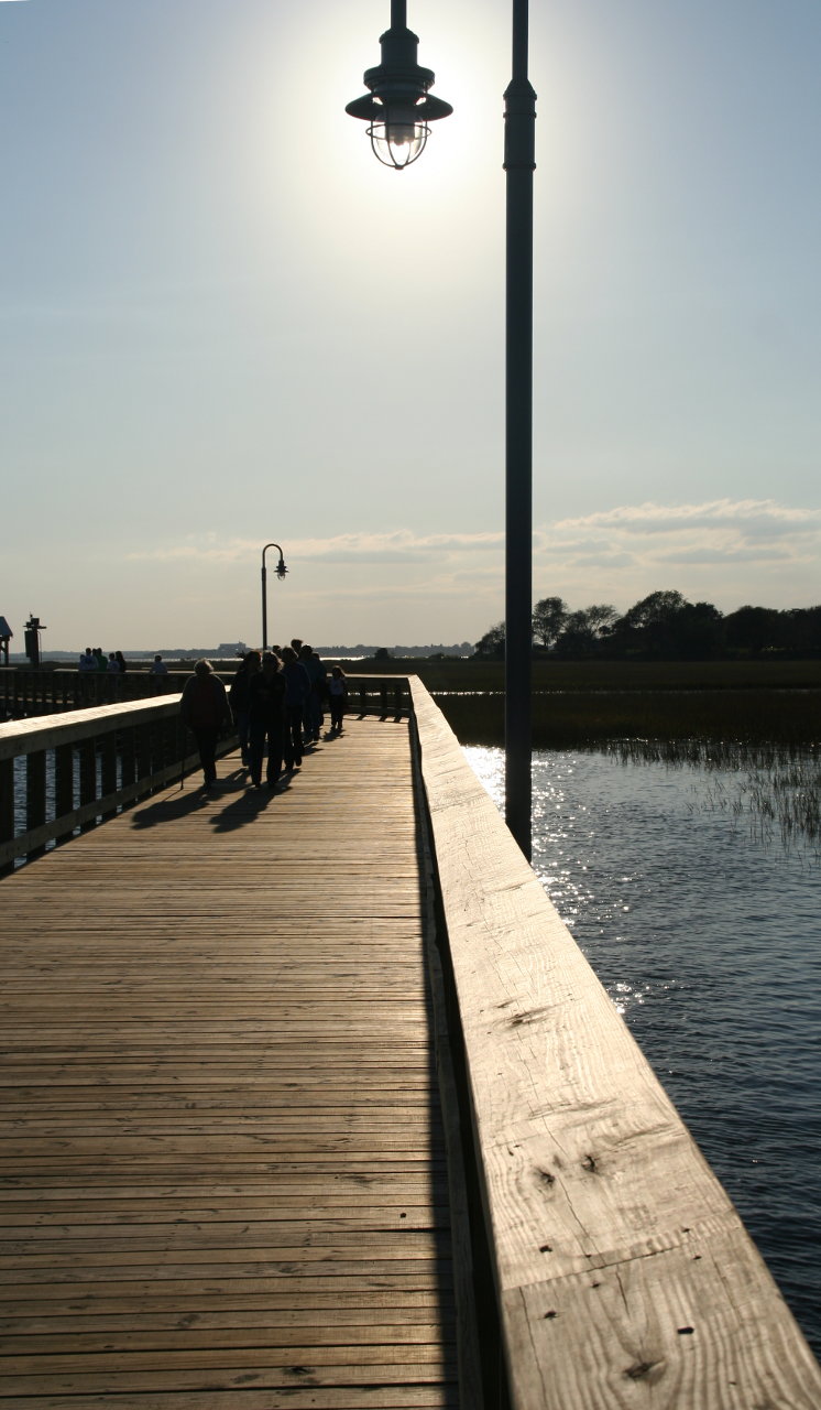 Silhouette of a street lamp against the sun.  The street lamp is attached to a boardwalk over the marsh.