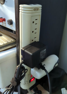 A power strip mounted to the side of a bookshelf with a 5V wall wart power supply and flashlight plugged in
